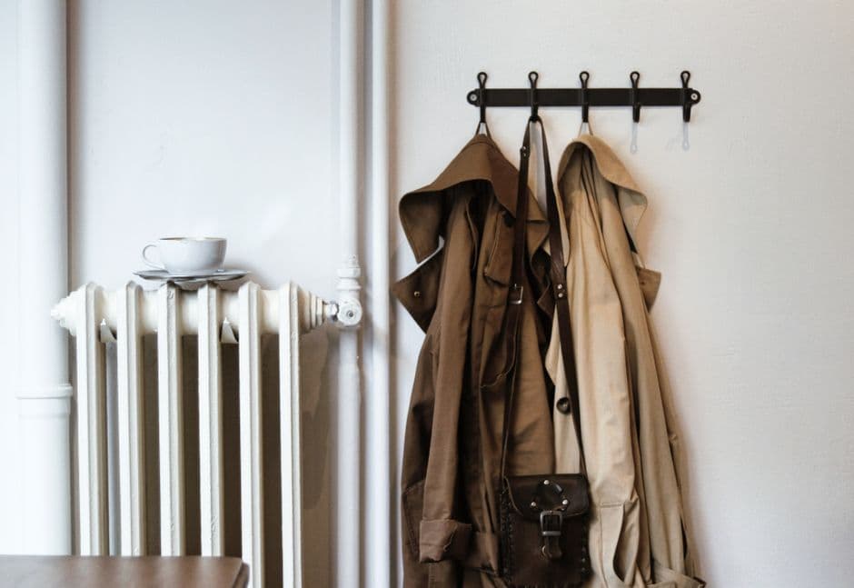 Two coats and a leather bag overlapping on a five-hook wall rail beside a radiator